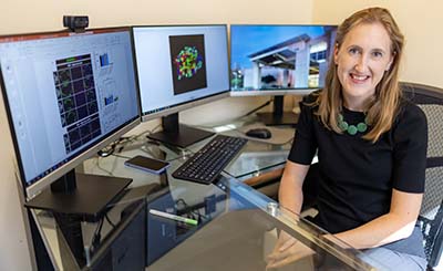 Associate Professor Bethany Cummings sits at her desk, working at her computer, which includes three monitors. 