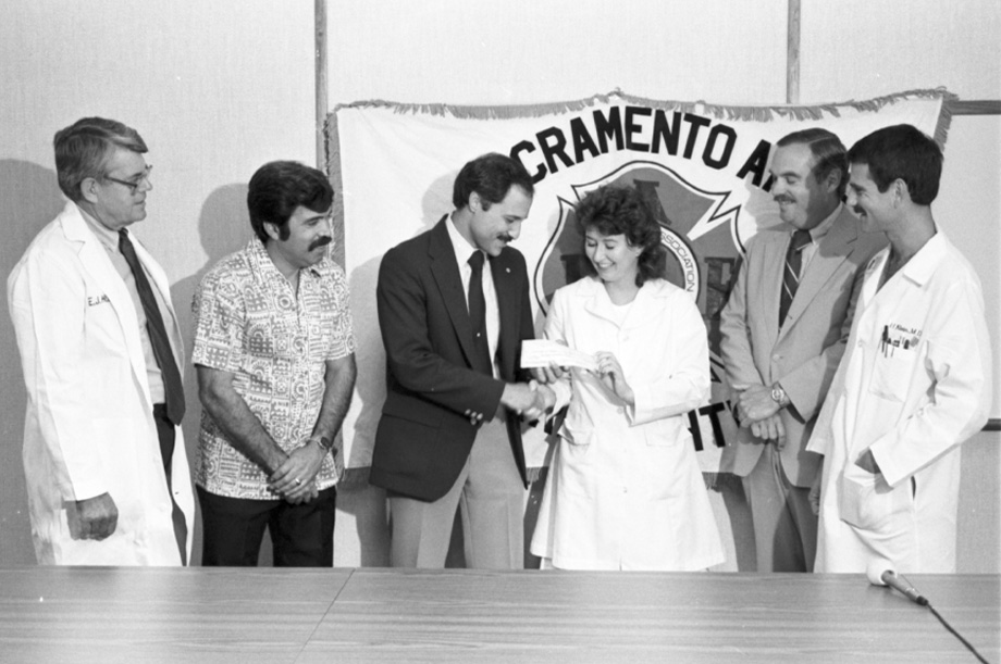 Five people standing against wall as a man, third from left, hands check to health care worker in white coat