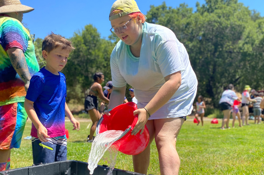 Rayne Carr pours bucket of water into larger tub while Everett Fifth-Lince looks on.