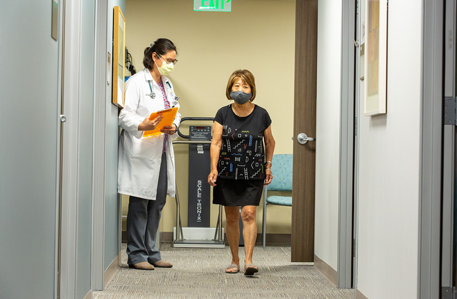 Woman wearing dress walks while clinician in white lab coat writes assessment on a clipboard.