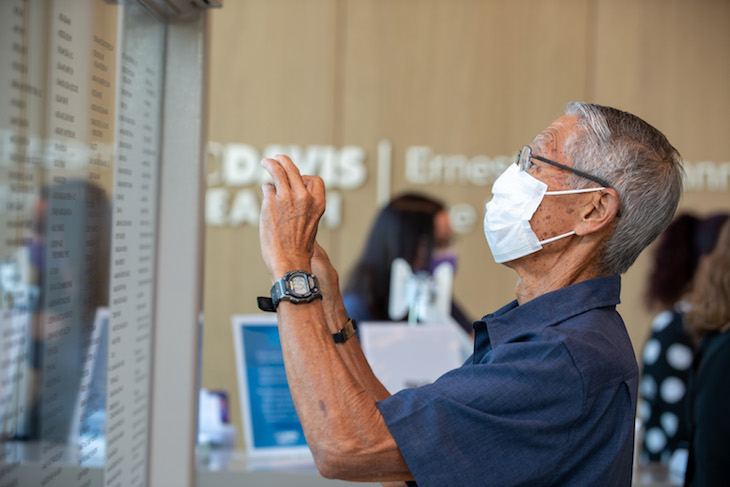 A man wearing a mask takes a photo with his phone of glass display containing names. 