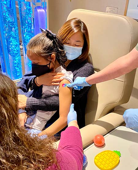 A woman sits in a medical chair, holding her adolescent daughter in the “comfort hold” while a nurse places a rainbow-colored bandage on the girl’s arm. 