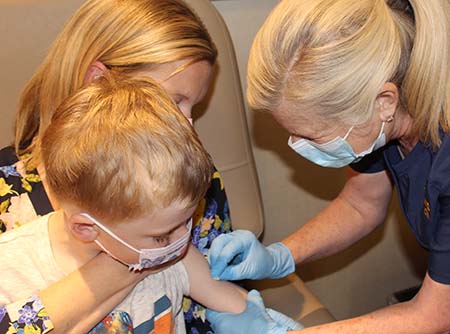 A woman sits in a medical chair, holding her young son in the “comfort hold” while he a nurse cleans his arm with a sanitary wipe in preparation for a shot.