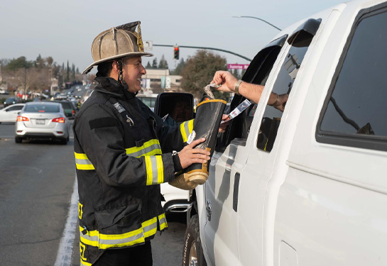 Firefighter in full gear stands outside a white truck holding fire boot while driver places dollar bill into boot