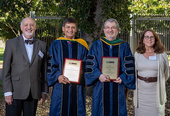 Three men and one woman stand in front of a shady tree; the two men in the middle are in academic robes and hold placards. 