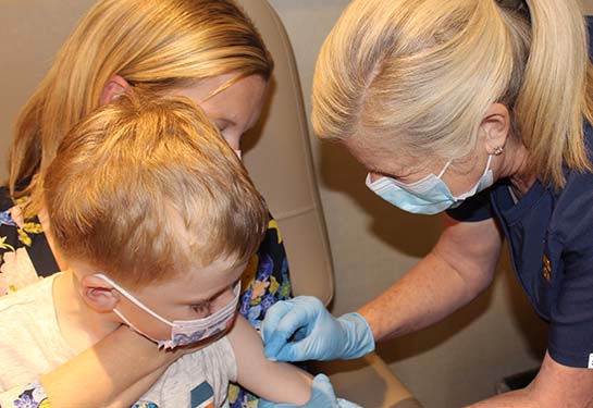 A woman sits in a medical chair, holding her young son in the &#x201c;comfort hold&#x201d; while he a nurse cleans his arm in preparation for a shot