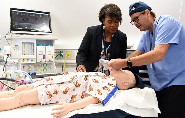 A man and a woman are holding a tube in the mouth of a child manikin with medical equipment in the background.