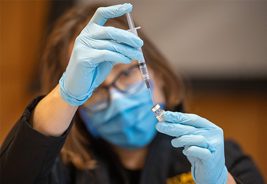 A UC Davis Health employee wearing blue gloves and blue face mask uses a syringe to extract vaccine from a small bottle 