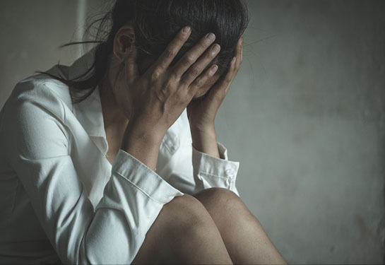 Woman with long brown hair wearing a white shirt sitting with head in hands and knees pulled to chest