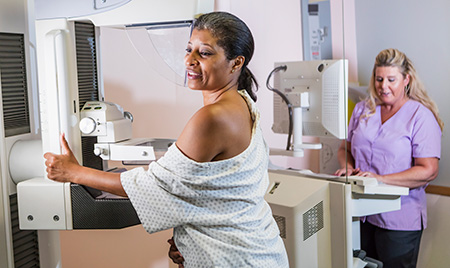 A mature African American woman in her 40s wearing a hospital gown, getting her mammogram  being helped by a technologist, a blond woman wearing scrubs.