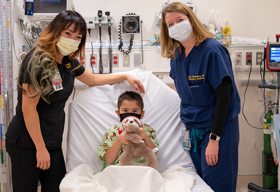 Young male holding a teddy bear a nurse on each side of the bed