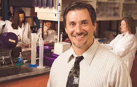 Dr. Murphy in his lab smiling to the camera. Behind him three individuals in white gowns 