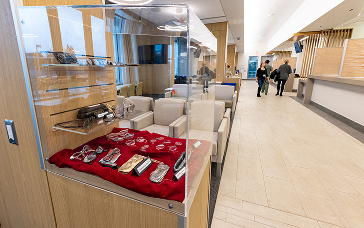 A display case with a red cloth and vintage eyeglasses is next to a waiting area with chairs and a long hallway with beige tile. 