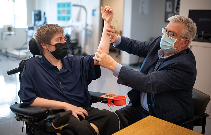 A young man with Duchenne muscular dystrophy is sitting in a wheelchair. A doctor is folding the arm of the man and pressing at the elbow