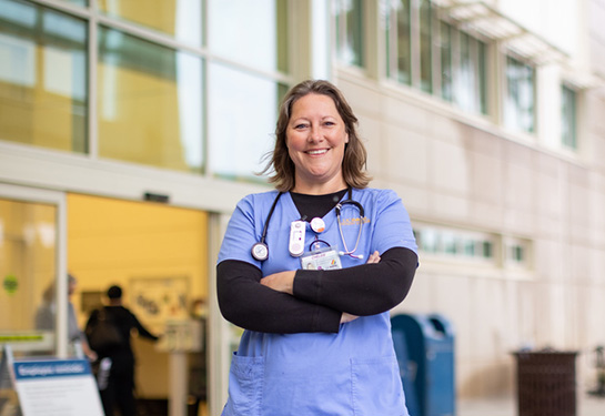 Chelsie Gilbeau wearing blue scrubs standing front of hospital with arms crossed