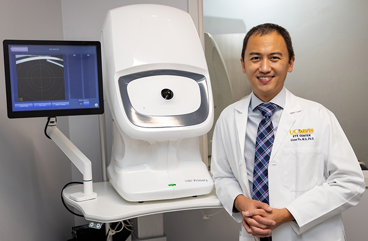 A physician in a white coat stands next to a white machine with a dark camera hole in the middle. A computer monitor is next to the machine showing a blue screen. 