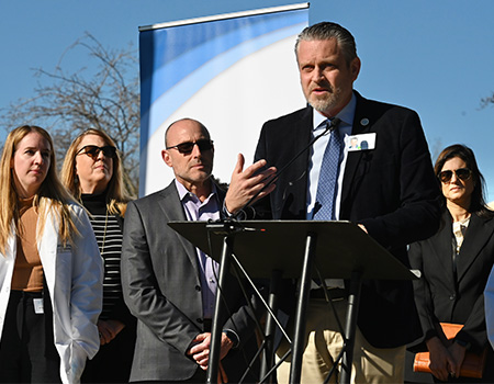 Jonathan Porteus, CEO of WellSpace Health, stands at the podium and speaks into microphone, surrounded by several health professionals 