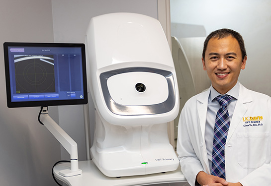 A physician in a white coat stands next to a white machine with a dark camera hole in the middle. A computer monitor is showing a blue screen.