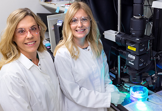 Two women with blond hair and in white lab coats and safety glasses stand in a lab next to a black imaging device with a glass dish underneath