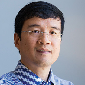 A man with short dark hair and wire-rimmed glasses wearing a blue striped shirt and blue patterned tie faces the camera with a half-smile. 