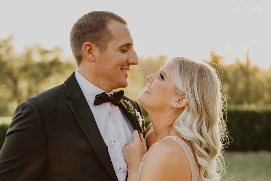 Man, left, in tuxedo smiles and hugs his wife, right, as she smiles at him on their wedding day