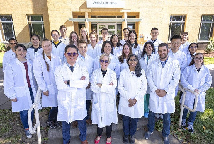 A group of scientists dressed in the white lab coats standing in front of a building. Dr. Wang and Dr. Farmer are standing in the front row.