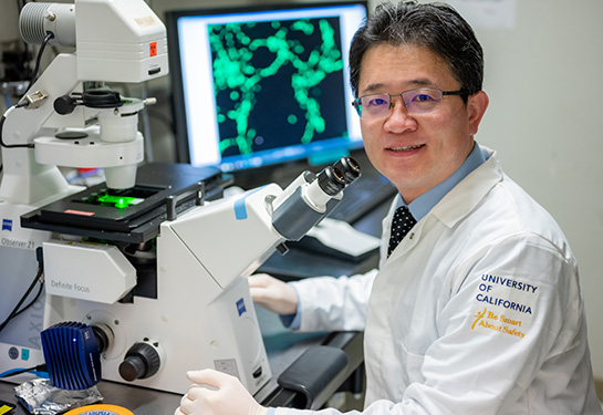 Researcher in white lab coat with a microscope and computer on the desk