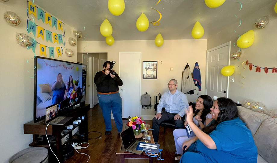 Two female medical students and their husbands watch a TV screen displaying a Zoom call where students shared results of Match Day