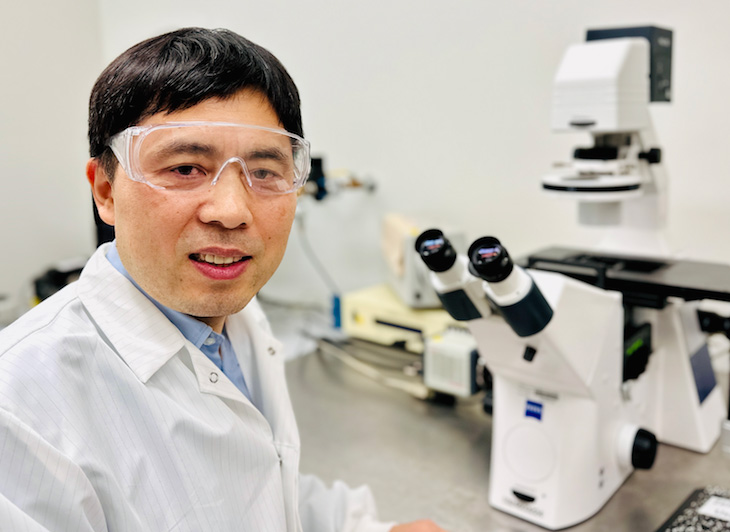 A man with dark hair wearing a white lab coat and lab goggles sits in front of a white microscope. 