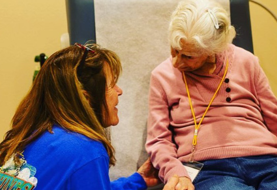 young woman knees beside older woman in exam room chair and holds her arm