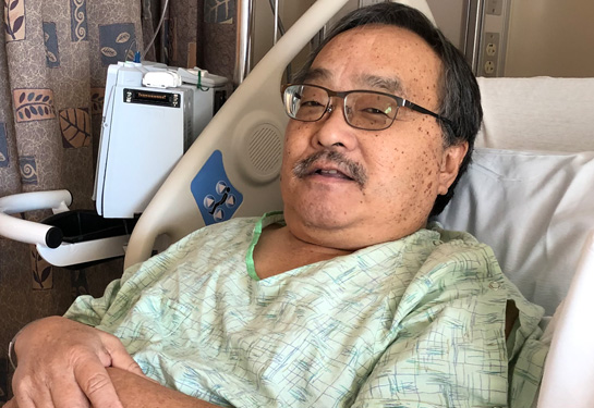 An older man wearing glasses and a green and white hospital gown lies in a hospital bed smiling