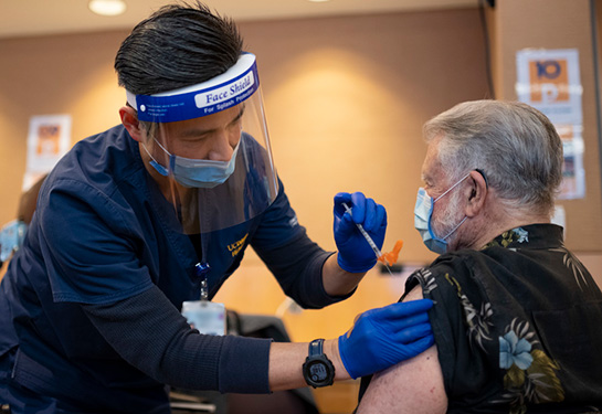 Nurse wearing mask and face shield injecting vaccination in person arm with sleeve rolled up. 