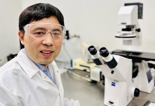 A man with dark hair wearing a white lab coat and lab goggles sits in front of a white microscope. 