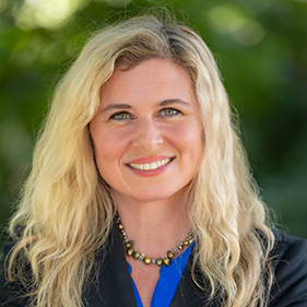 A formal headshot of a woman with blond hair wearing a dark blazer and a bright blue shirt.