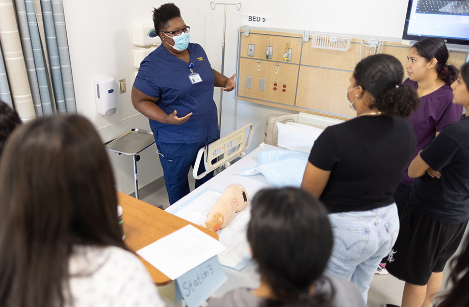 Nurse wearing mask and scrubs stands behind hospital bed talking with high school students gathered nearby around the bed.