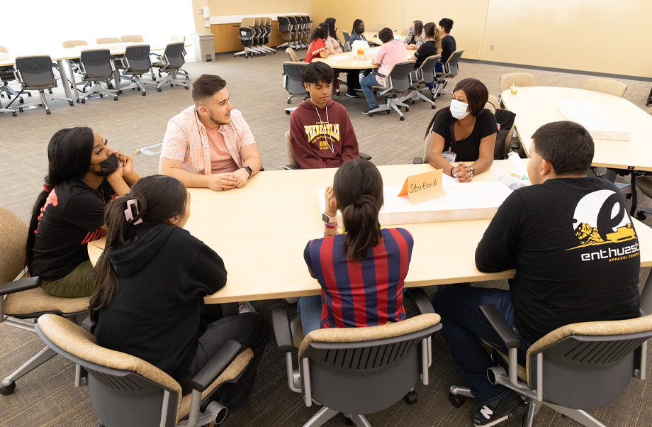 Students sit around a propeller-shaped table talking to one another.