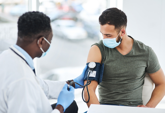 A doctor in a white coat and surgical mask holds a gloved hand on a blood pressure cuff that&#x2019;s wrapped around the left arm of a young man