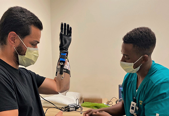 Two men wearing masks face each other, man on left has a prosthetic left hand. 