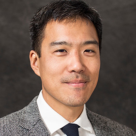 Man with dark hair wearing a white shirt, gray blazer over gray background.