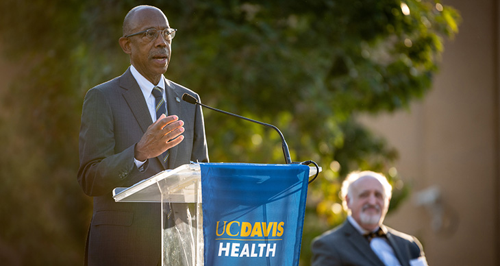 A man in a grey suit stands at a podium giving a speech in an outdoor setting. 