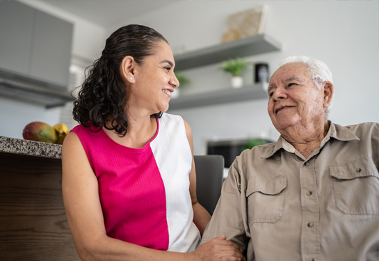 Daughter, left, sits on sofa next to older father smiling and holding his arm