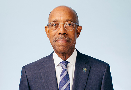 A man in a blue suite and a blue striped tie and white shirt smiles looking directly at the camera. 