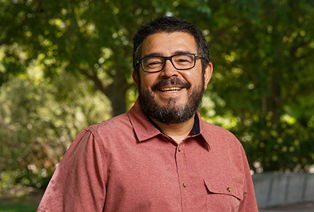 Latino man in orange shirt standing on grass under tree canopy