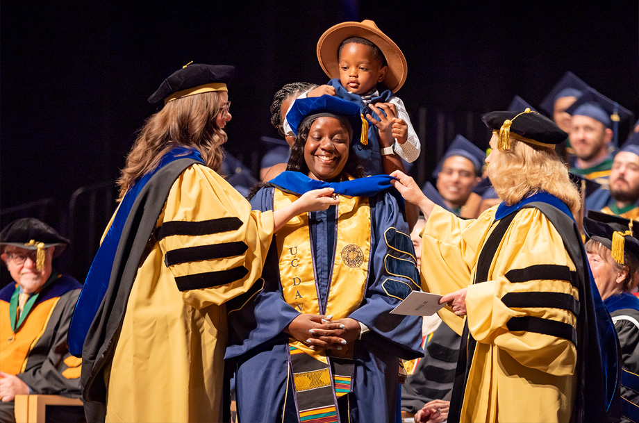 woman kneels during graduation as son hoods her from behind, two other faculty flank her