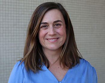 Smiling individual with shoulder-length brown hair, wearing light blue shirt.