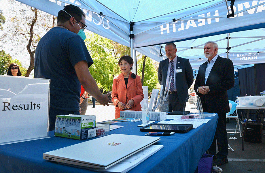 A man on left wearing teal face mask converses under a blue pop-up tent with a woman in orange suit and two men in blue suits