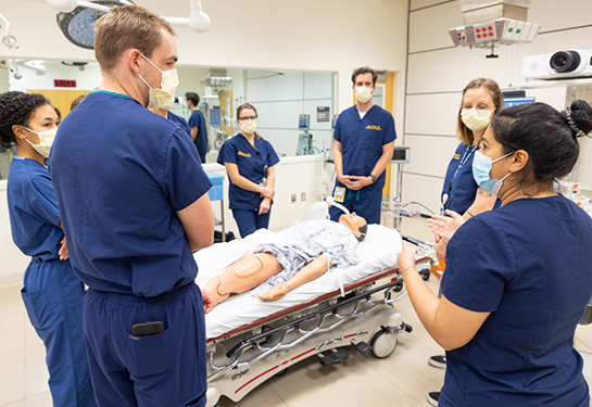 Group wearing blue scrubs surrounding bed with mannequin.