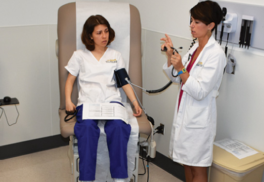 Nursing instructor, right, holds stethoscope in hand while student sits in patient chair with blood pressure cuff on arm