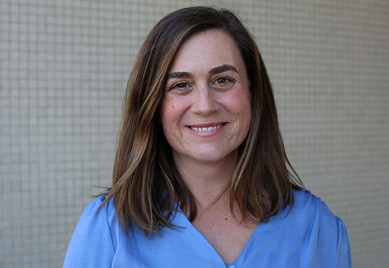 Smiling individual with shoulder-length brown hair, wearing light blue shirt.