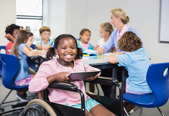 An elementary-aged African American girl in a wheelchair, wearing a pink shirt and blue shorts and holding a tablet, sits in front of a table